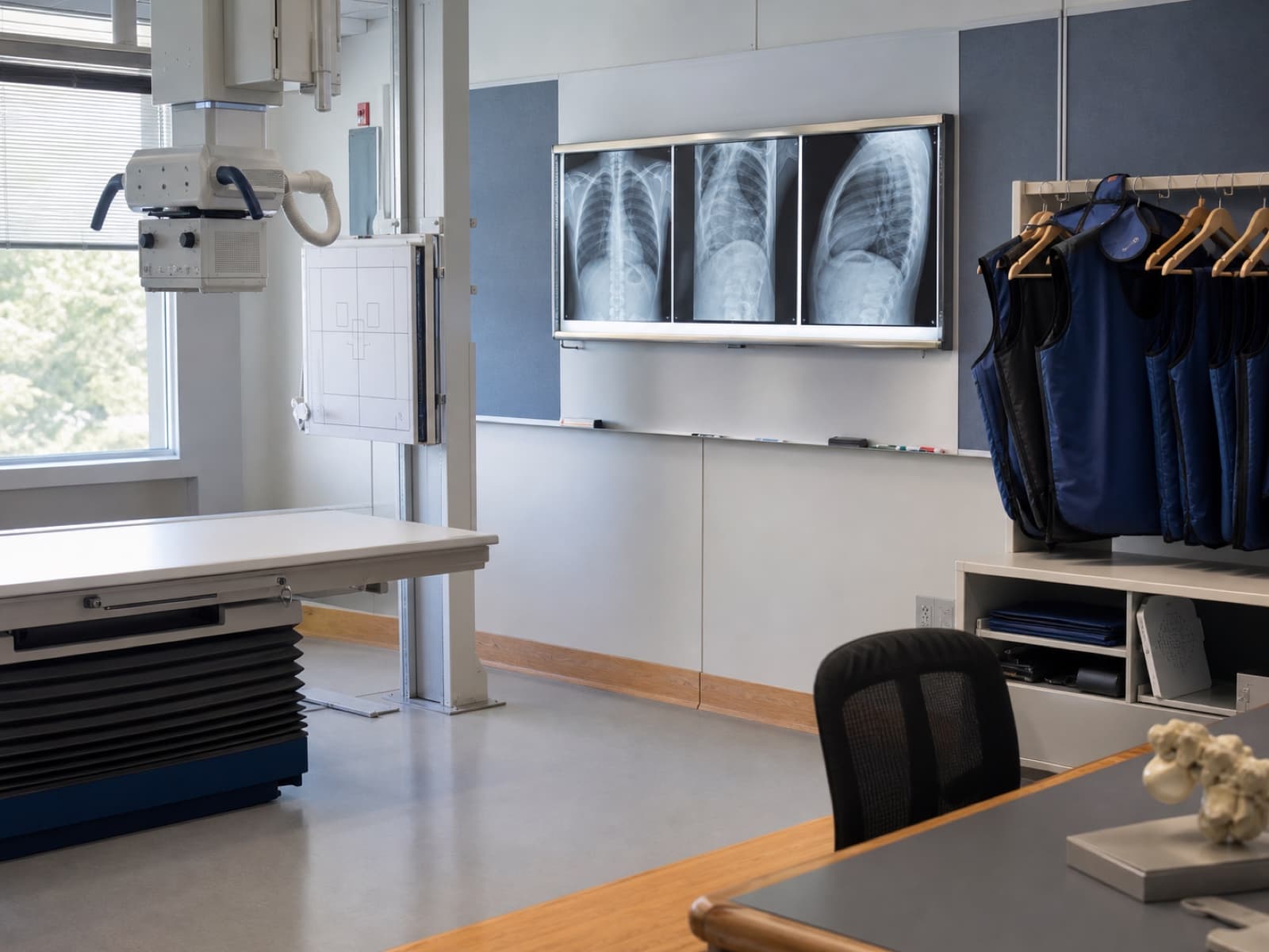 Sala de radiologia didática com mesa de exames, negatoscópio e aventais plumbíferos.